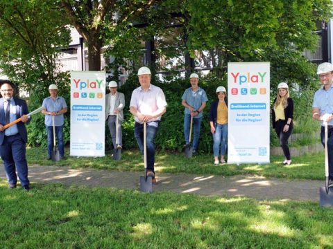 Gruppe Personen mit Spaten und Bauhelmen stehen vor Bäumen auf Wiese mit Yplay-Bannern, Glasfaserausbau Niddatal Kaichen