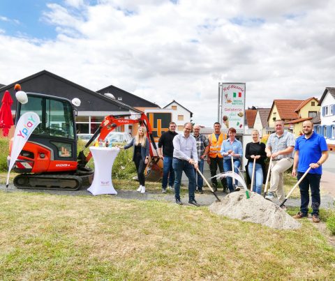 Gruppe von Personen stehen vor Sandhaufen mit Bauhelmen und Spaten neben Bagger und Yplay Fahne zum Spatentisch in Niddatal-Ilbenstadt