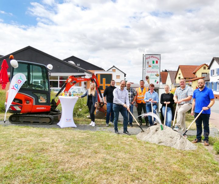 Gruppe von Personen stehen vor Sandhaufen mit Bauhelmen und Spaten neben Bagger und Yplay Fahne zum Spatentisch in Niddatal-Ilbenstadt